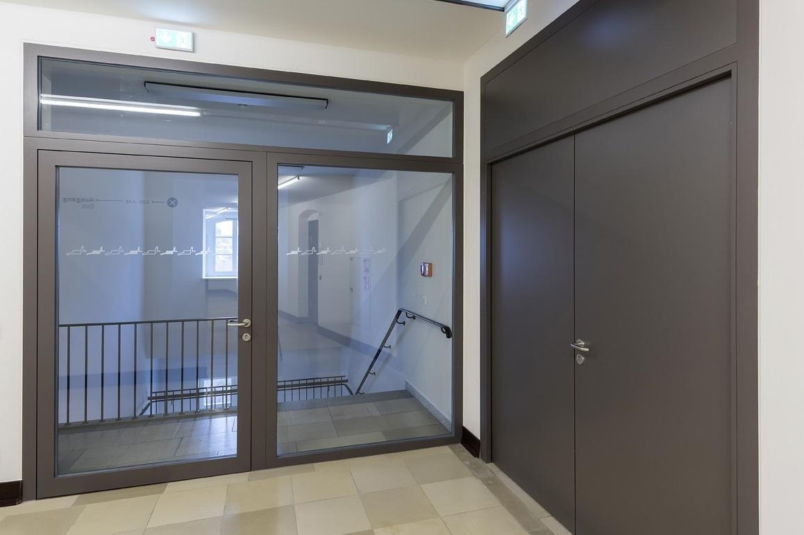 Durchsichtige Glast&uuml;r mit modernem Rahmen, Zugang zu einem Raum mit Treppe und Gel&auml;nder im Hintergrund.