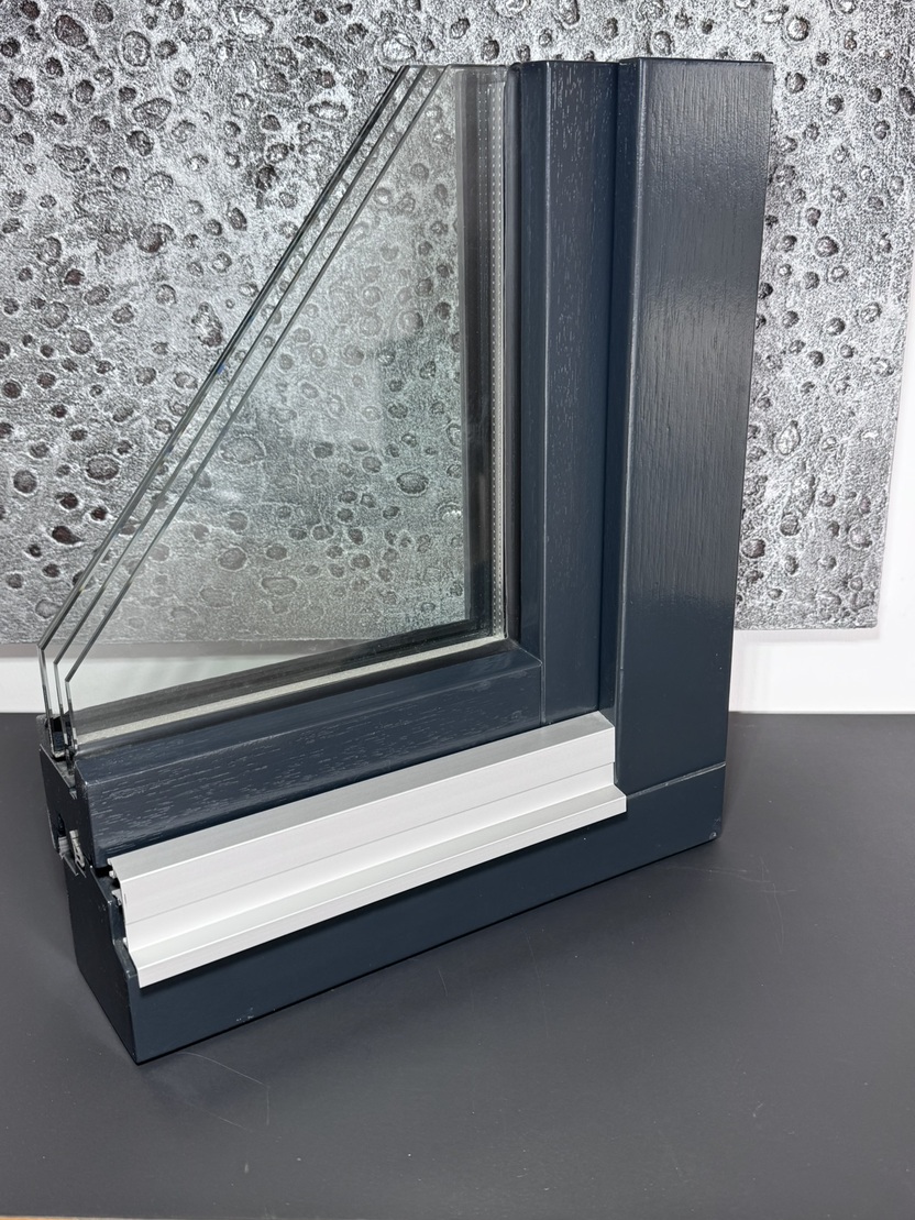 Detailaufnahme eines Fensterprofils mit Glas und Aluminiumrahmen vor einer strukturierten grauen Wand.