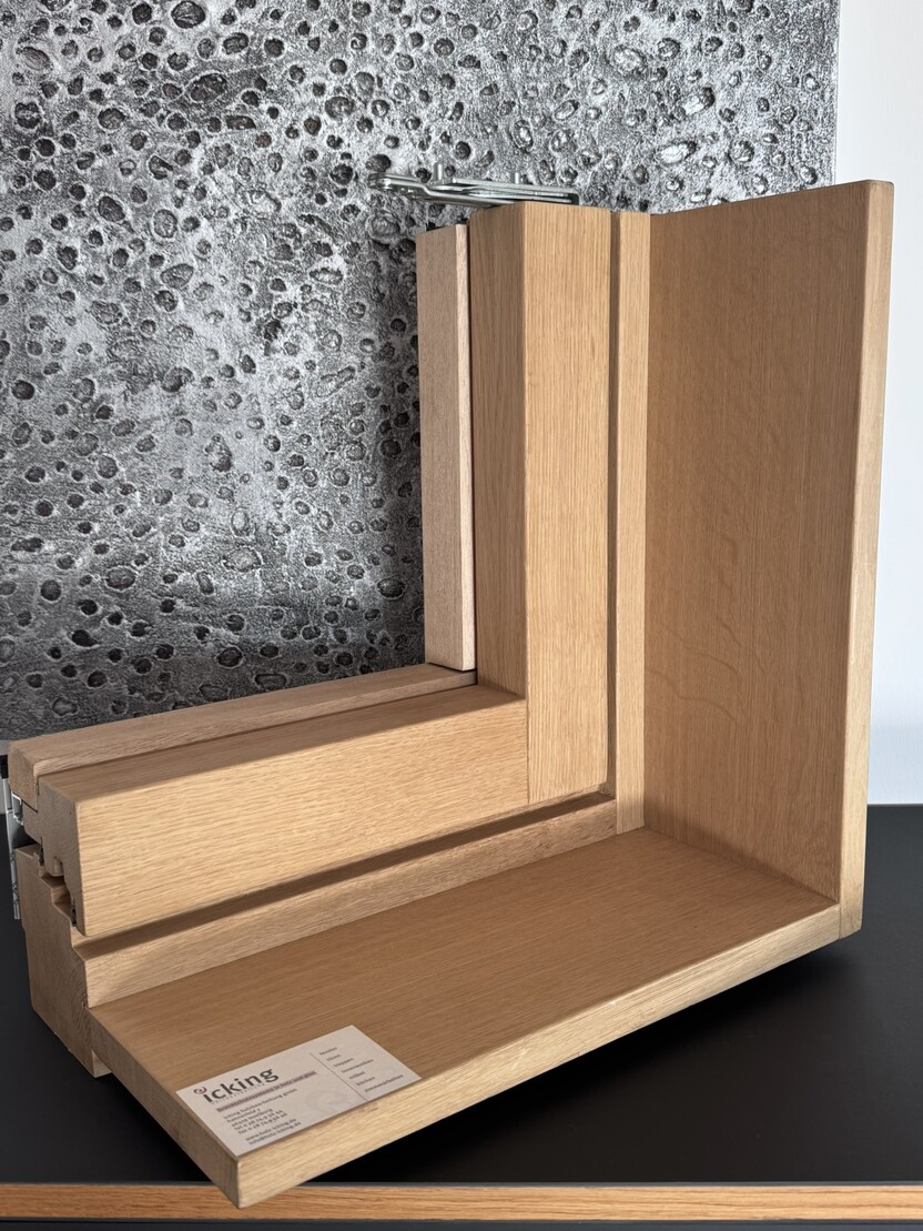 Detailaufnahme eines h&ouml;lzernen Fensterrahmens in L-Form mit einer grauen, strukturierten Wand im Hintergrund.