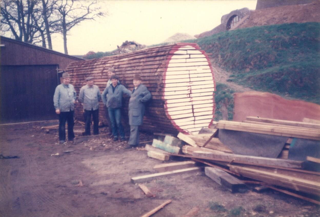 F&uuml;nf Personen stehen neben einem gro&szlig;en Baumstamm, umgeben von Holzresten und einem landwirtschaftlichen Hintergrund.