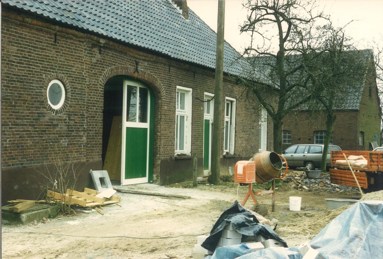 Baustelle mit einem Haus im Hintergrund, Mauerwerksfassade, gr&uuml;nen T&uuml;ren, Baum, Baustellenmaterialien und einer Betontrommel.