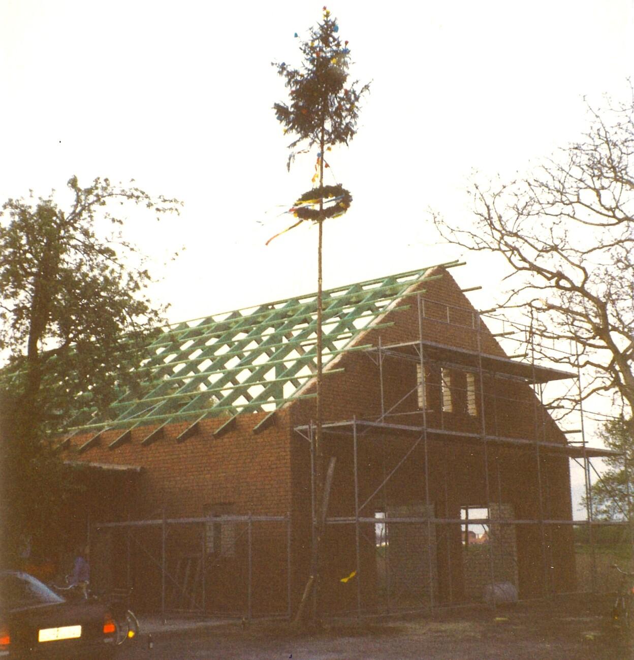 Neubau eines Hauses mit aufragendem Baum am Dach und Ger&uuml;stkonstruktion, umgeben von B&auml;umen und Fahrzeugen.