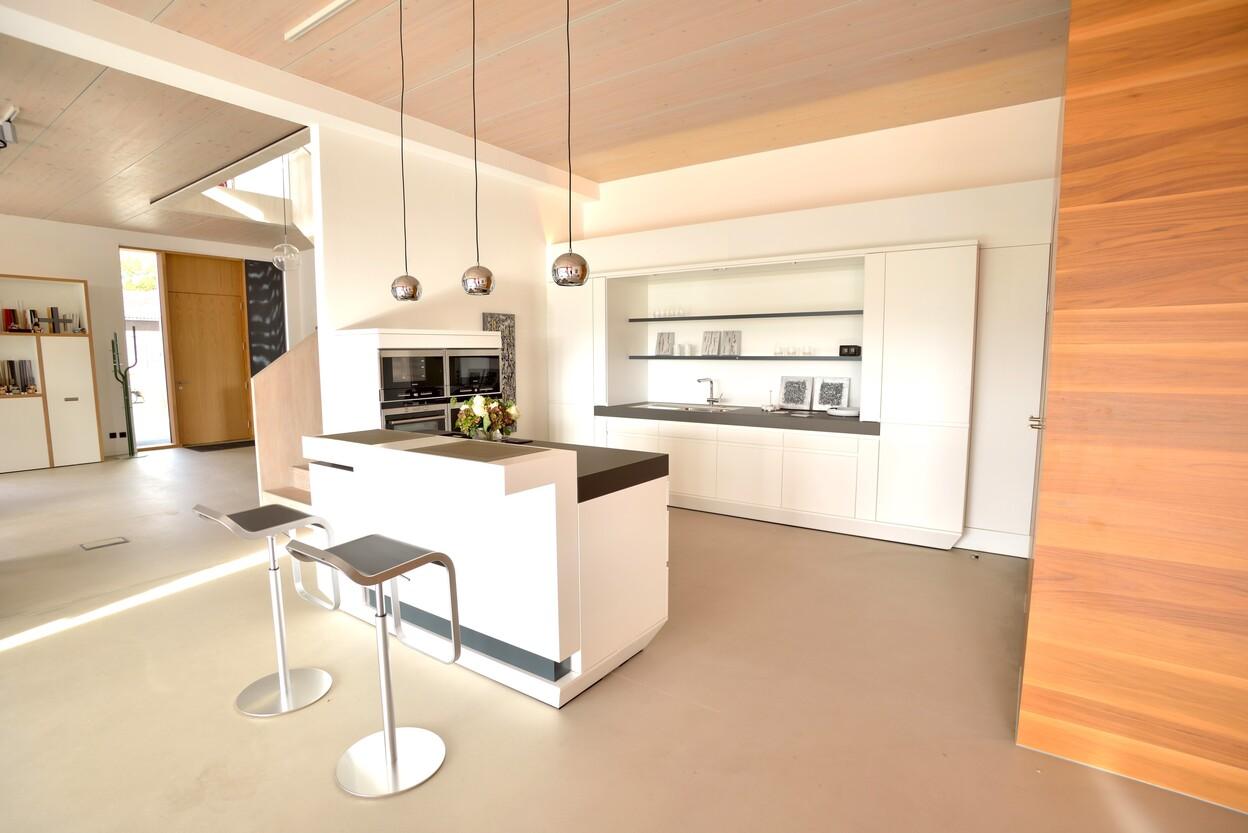 Moderne, helle K&uuml;che mit wei&szlig;en M&ouml;beln, K&uuml;chentresen und St&uuml;hlen sowie Holzdetails in warmen T&ouml;nen.