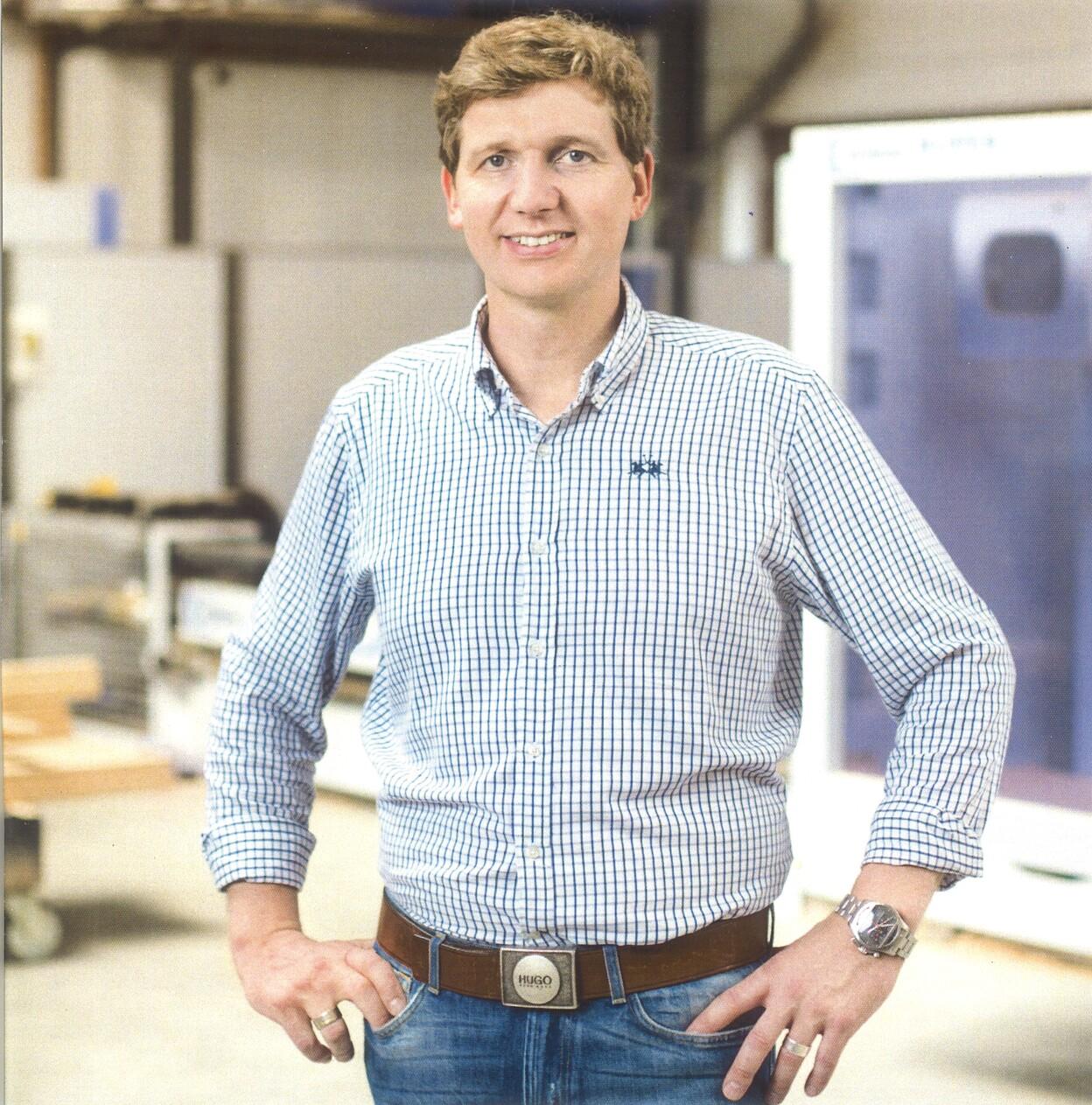 Mann mit kariertem Hemd und G&uuml;rtel steht in einer Werkstatt, im Hintergrund Maschinen und Holzmaterialien sichtbar.