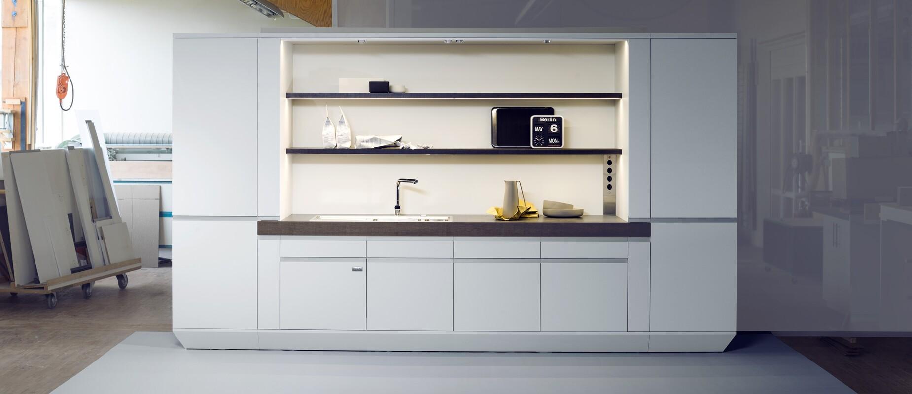 Moderne, wei&szlig;e K&uuml;chenzeile mit Sp&uuml;le, Regalen und Elektroger&auml;ten in einem Werkstattumfeld.