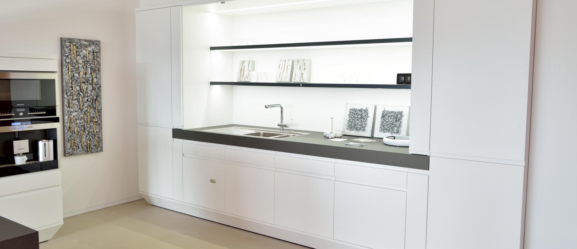 Moderne wei&szlig;e K&uuml;che mit schwarzer Arbeitsfl&auml;che, Sp&uuml;le und offenen Regalen, vor neutralem Hintergrund.