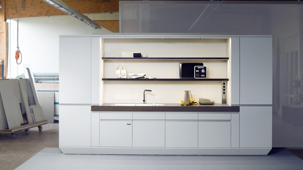 Moderne, wei&szlig;e K&uuml;chenzeile mit Sp&uuml;le, Regalen und Elektroger&auml;ten in einem Werkstattumfeld.