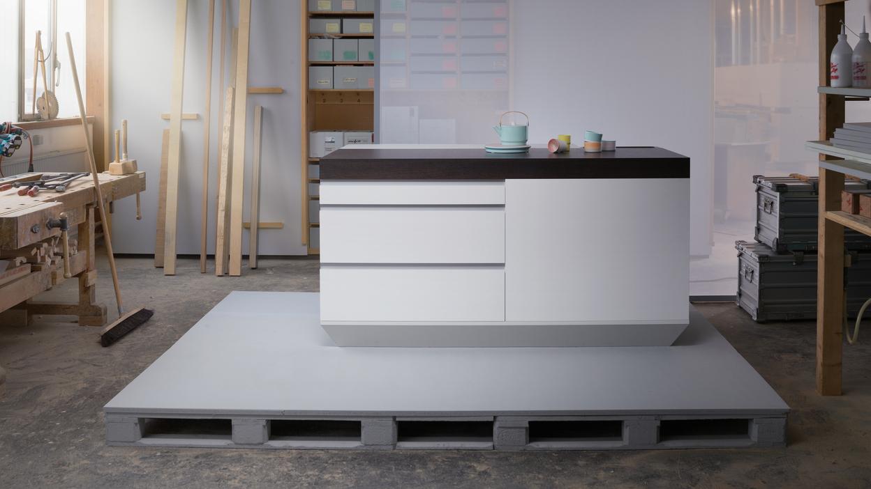 Moderne K&uuml;cheninsel mit Holzoberfl&auml;che, auf einem grauen Palettensockel in einem hellen Werkstattbereich.