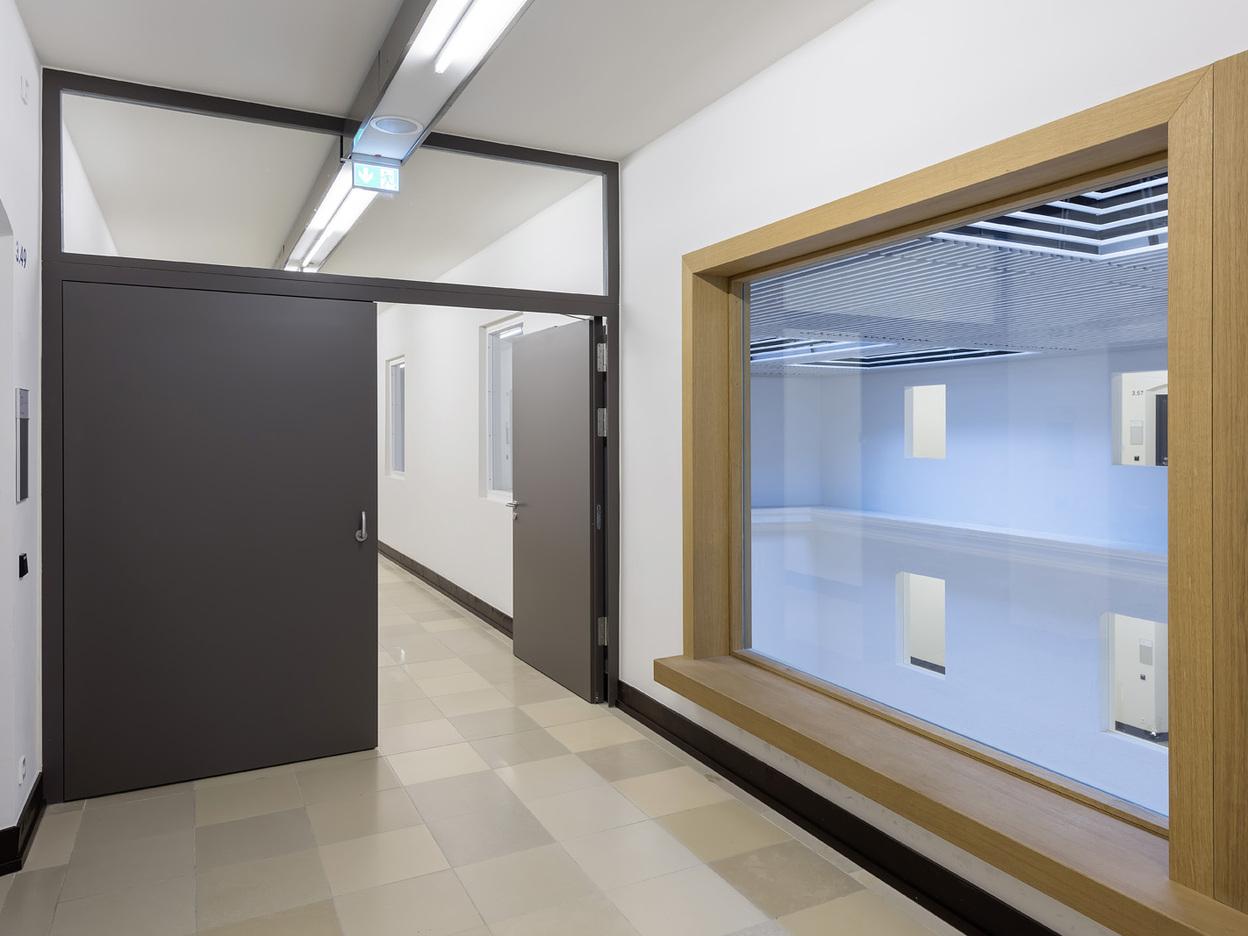 Blick in einen modernen Flur mit einer grauen T&uuml;r und einem gro&szlig;en Holzfenster, das einen blauen Raum zeigt.