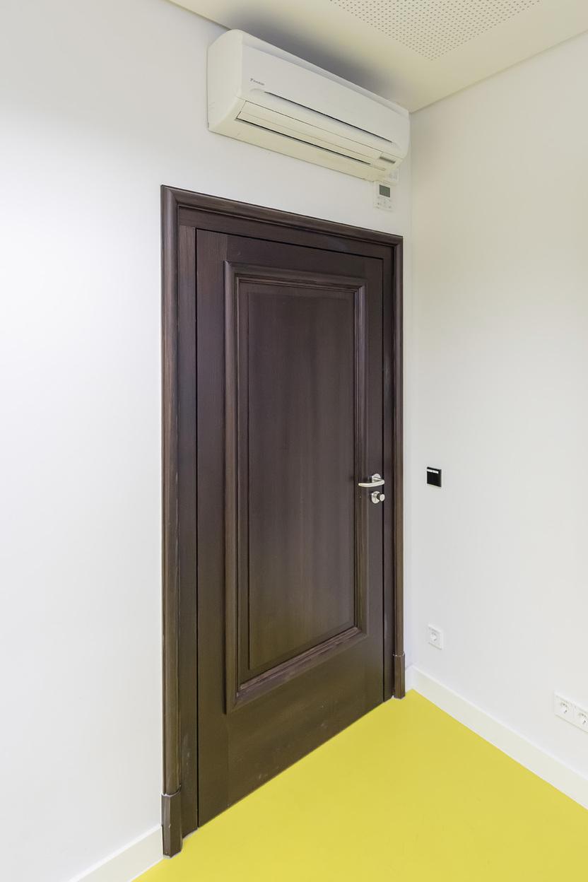 Braune T&uuml;r mit Griff in einem Raum mit wei&szlig;er Wand und Klimaanlage, gelbem Boden und Wandmontage f&uuml;r Fernseher.