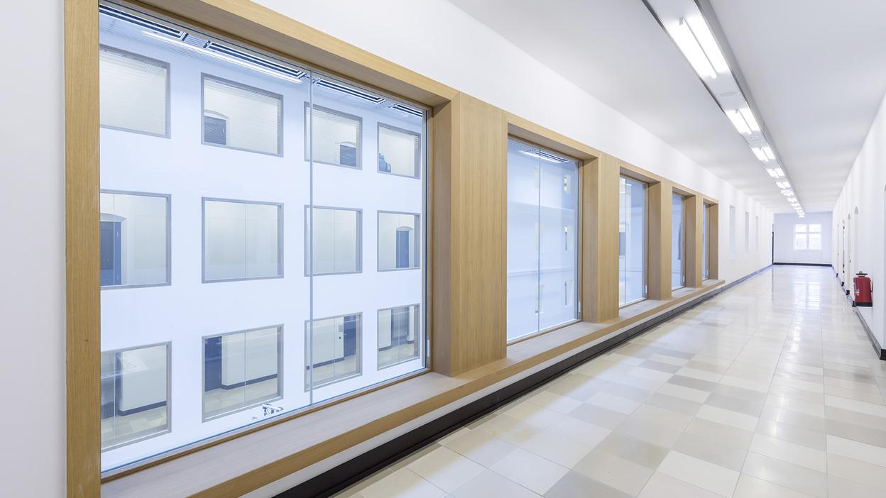 Langer, heller Flur mit gro&szlig;en Fenstern und Holzrahmen, der auf eine wei&szlig;e Au&szlig;enfassade blickt.
