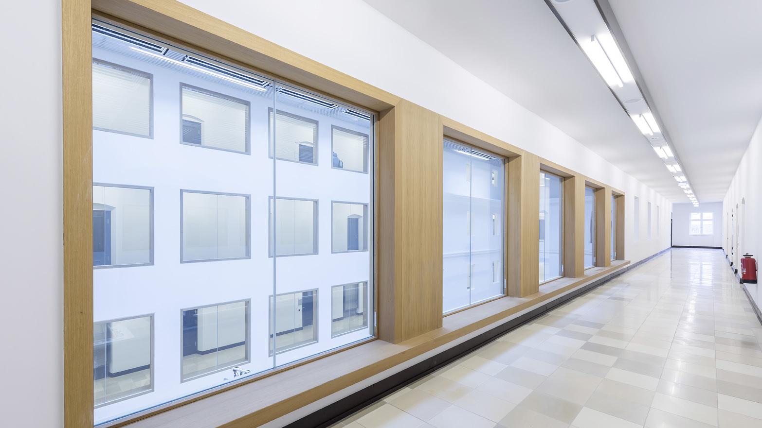 Langer, heller Flur mit gro&szlig;en Fenstern und Holzrahmen, der auf eine wei&szlig;e Au&szlig;enfassade blickt.
