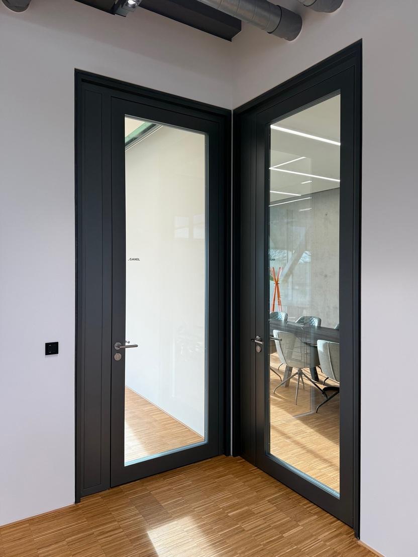 Zwei moderne Glast&uuml;ren in einem B&uuml;ro, eine steht offen und zeigt einen Besprechungsraum mit St&uuml;hlen und Tisch.