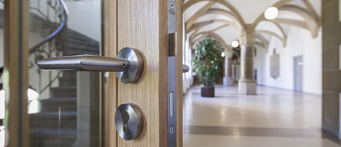 Detailaufnahme einer Holzt&uuml;r mit modernen Beschl&auml;gen, im Hintergrund ein heller, architektonisch gestalteter Flur.