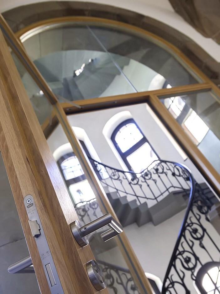 Holzt&uuml;r mit modernem T&uuml;rgriff, durchsichtigem Glas und Blick auf eine geschwungene Treppe im Hintergrund.