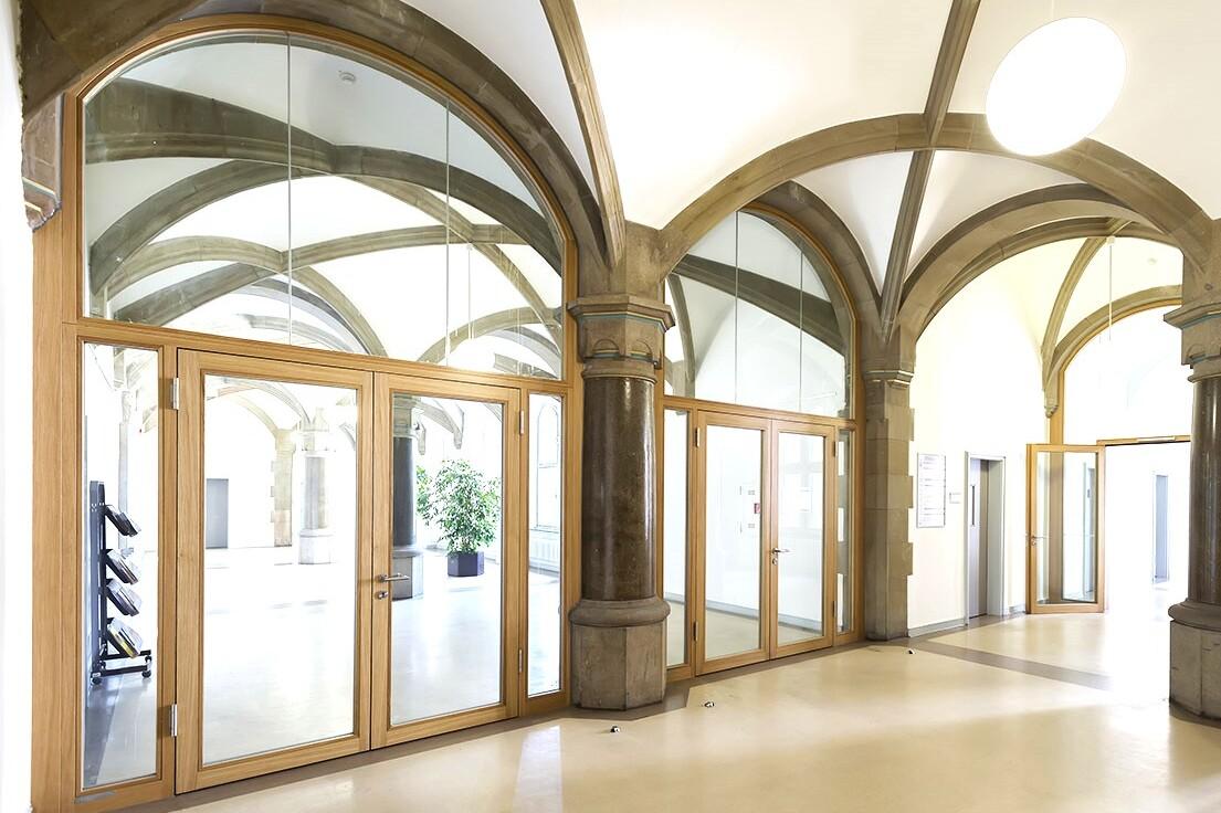 Lichtdurchfluteter Flur mit hohen Gew&ouml;lbedecken, Holz- und Glast&uuml;ren sowie einer gro&szlig;en Pflanze in der Ecke.