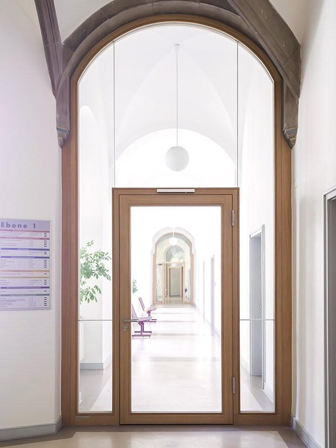 Holzt&uuml;r mit Glasf&uuml;llung, Blick auf einen langen, hellen Flur mit St&uuml;hlen und Pflanzen an den Seiten.