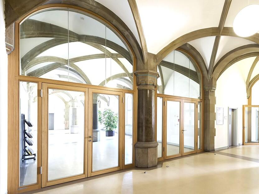 Lichtdurchfluteter Flur mit hohen Gew&ouml;lbedecken, Holz- und Glast&uuml;ren sowie einer gro&szlig;en Pflanze in der Ecke.