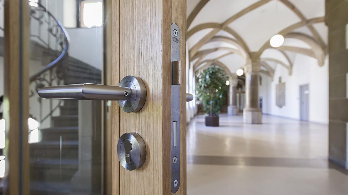 Detailaufnahme einer Holzt&uuml;r mit modernen Beschl&auml;gen, im Hintergrund ein heller, architektonisch gestalteter Flur.