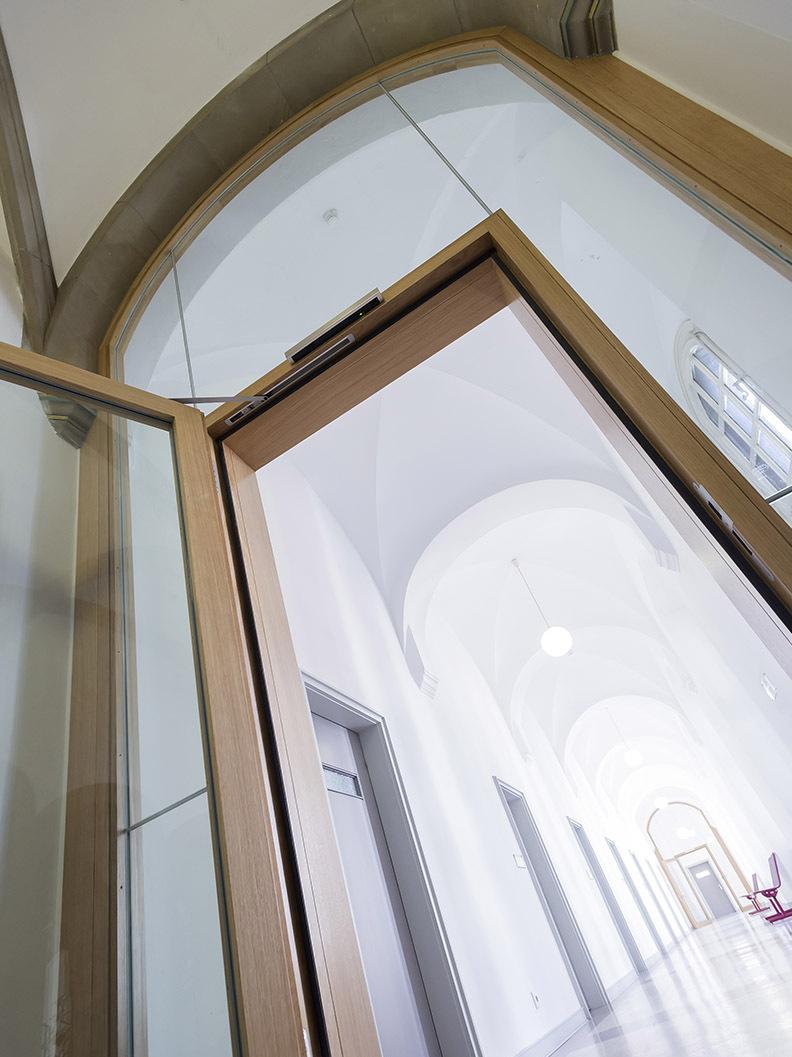 Durchsichtige Glast&uuml;r mit Holzrahmen &ouml;ffnet sich zu einem langen, hellen Flur mit mehreren T&uuml;ren und einer pinken Sitzgelegenheit.