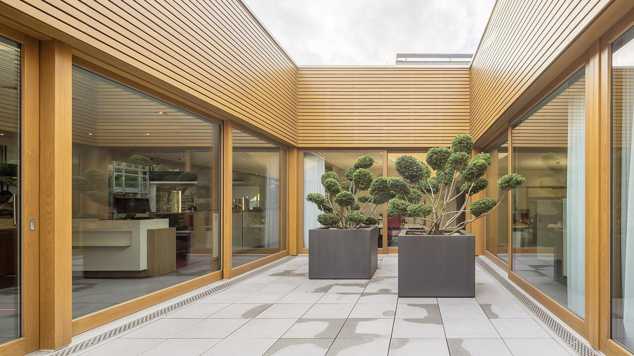 Innenhof mit Holzverkleidung und gro&szlig;en Fenstern, umgeben von zwei gro&szlig;en Pflanzk&uuml;beln mit Bonsai-B&auml;umen.