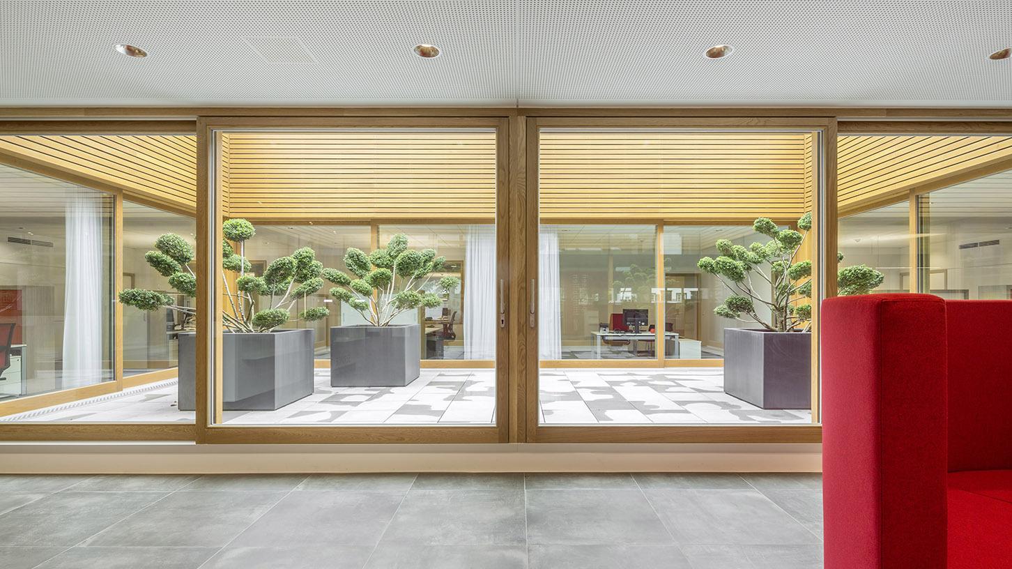 Blick auf eine moderne Glasfront mit Pflanzen in schwarzen K&uuml;beln und einem roten Sofa im Vordergrund.