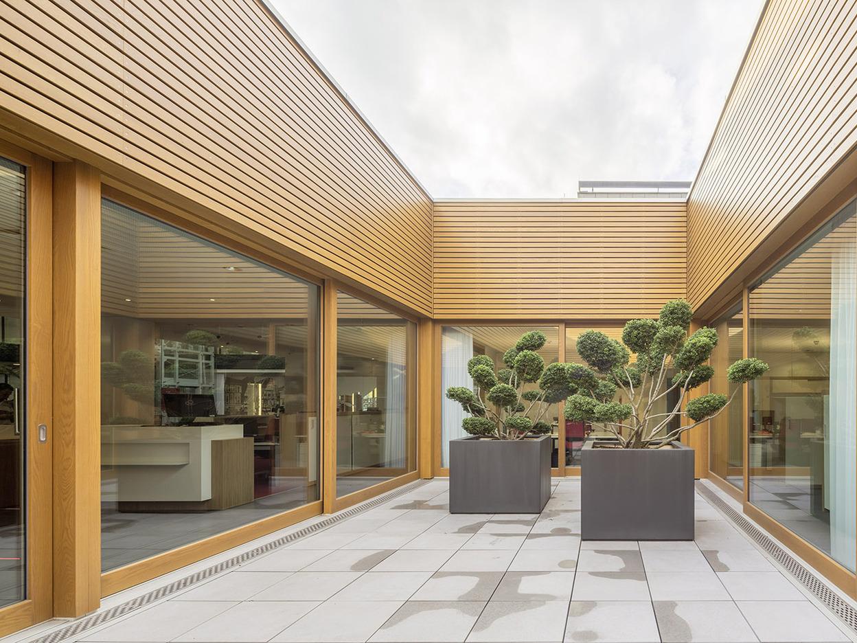 Innenhof mit Holzverkleidung und gro&szlig;en Fenstern, umgeben von zwei gro&szlig;en Pflanzk&uuml;beln mit Bonsai-B&auml;umen.