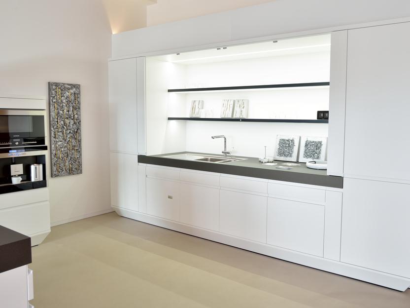 Moderne, minimalistische K&uuml;che mit wei&szlig;en Schr&auml;nken, schwarzer Arbeitsfl&auml;che und beleuchtetem Regal.