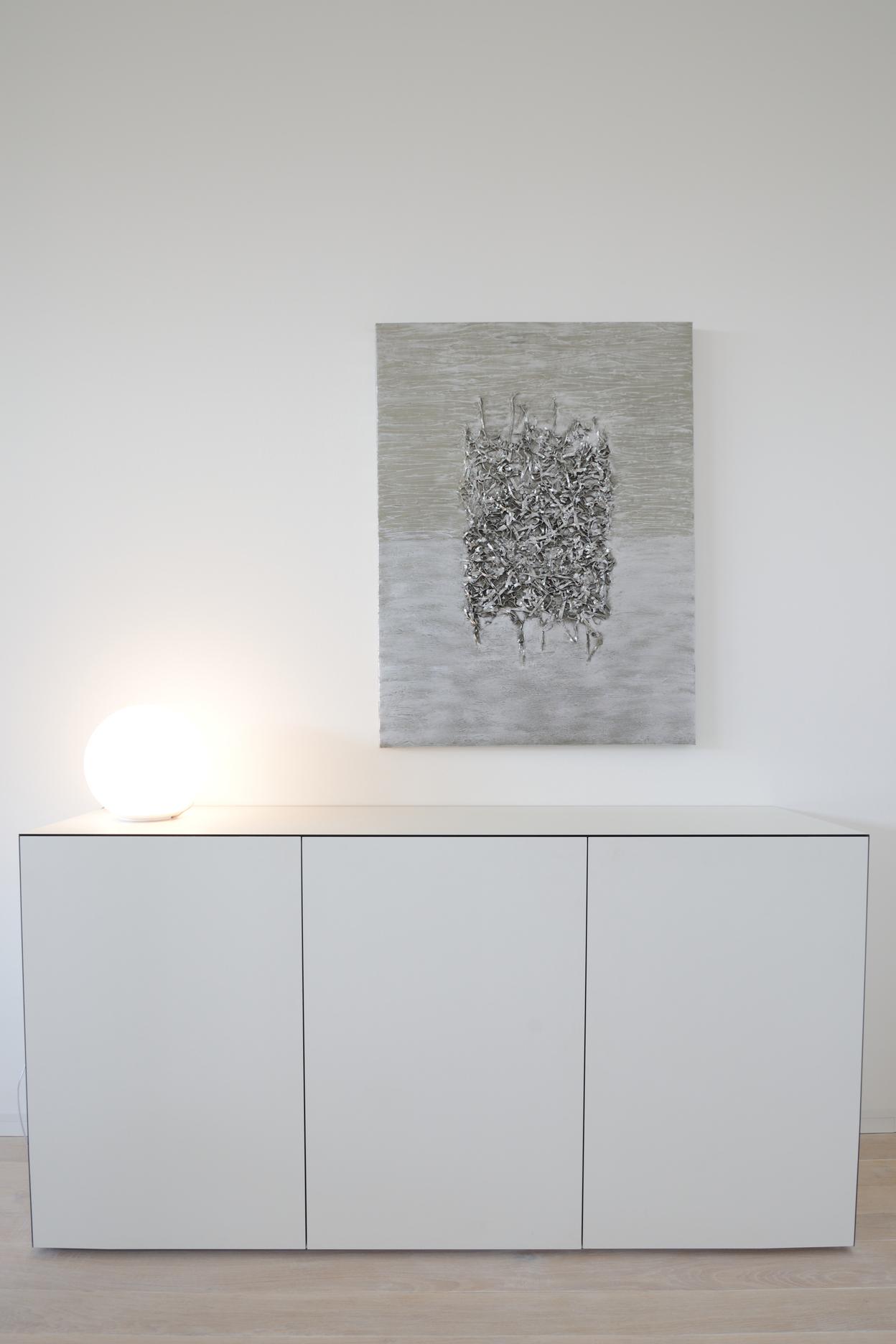 Wei&szlig;er Sideboard mit drei T&uuml;ren, beleuchtet von einer runden Lampe, im Hintergrund ein Wandbild in gedeckten Farben.