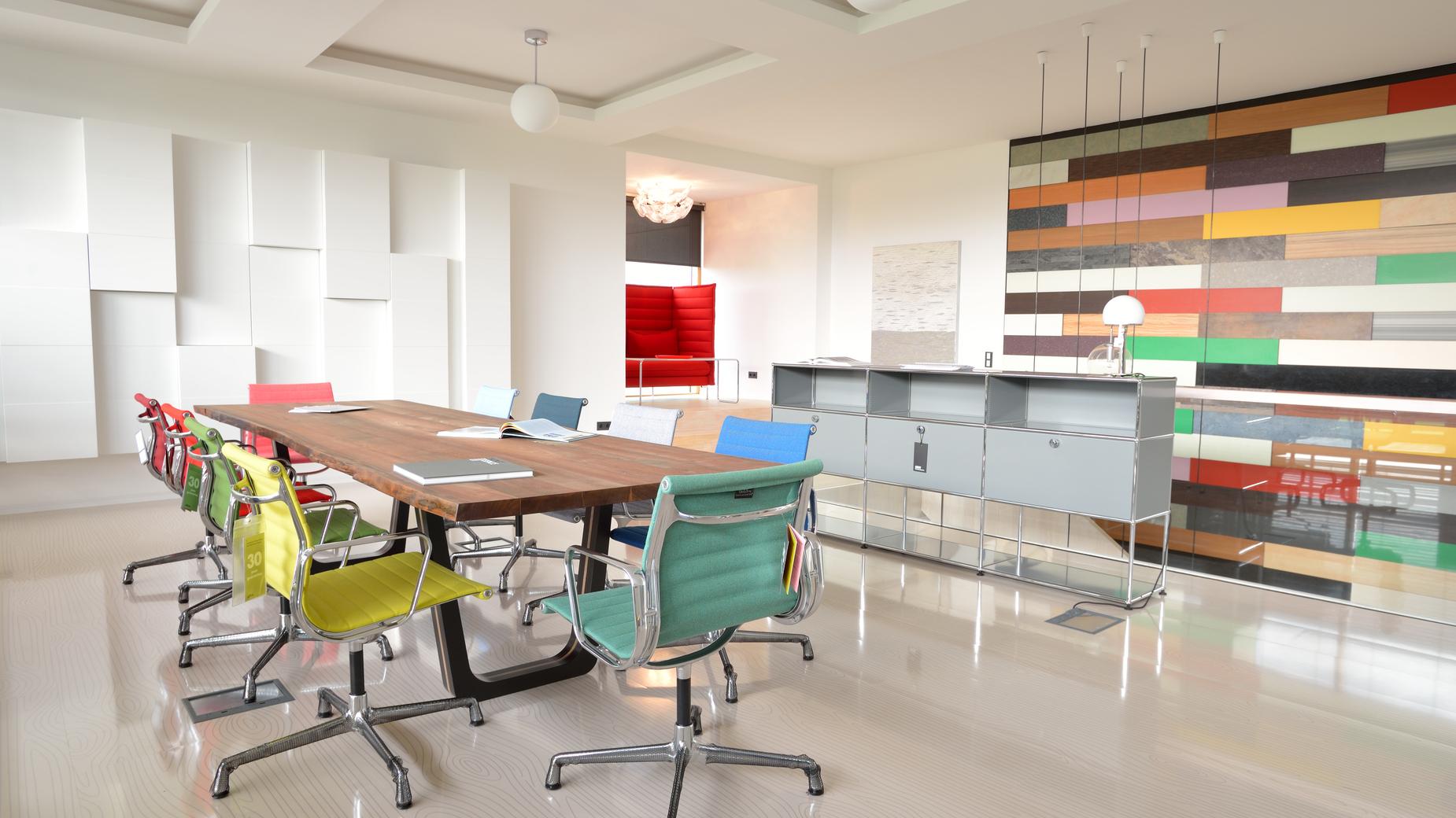 Modernes B&uuml;ro mit langem Holztisch, bunten St&uuml;hlen und einer farbigen Wandgestaltung im Hintergrund.