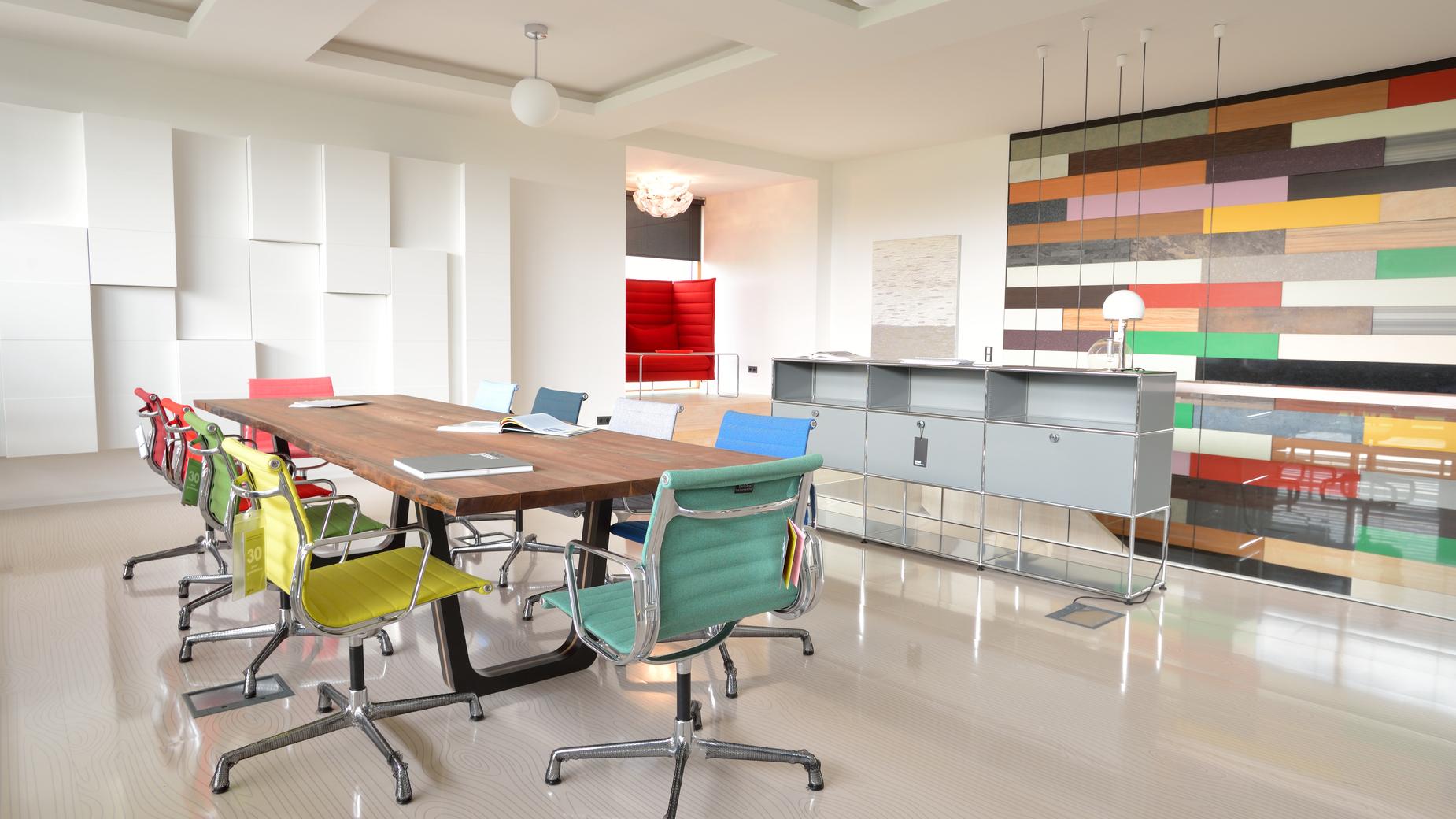Modernes B&uuml;ro mit langem Holztisch, bunten St&uuml;hlen und einer farbigen Wandgestaltung im Hintergrund.