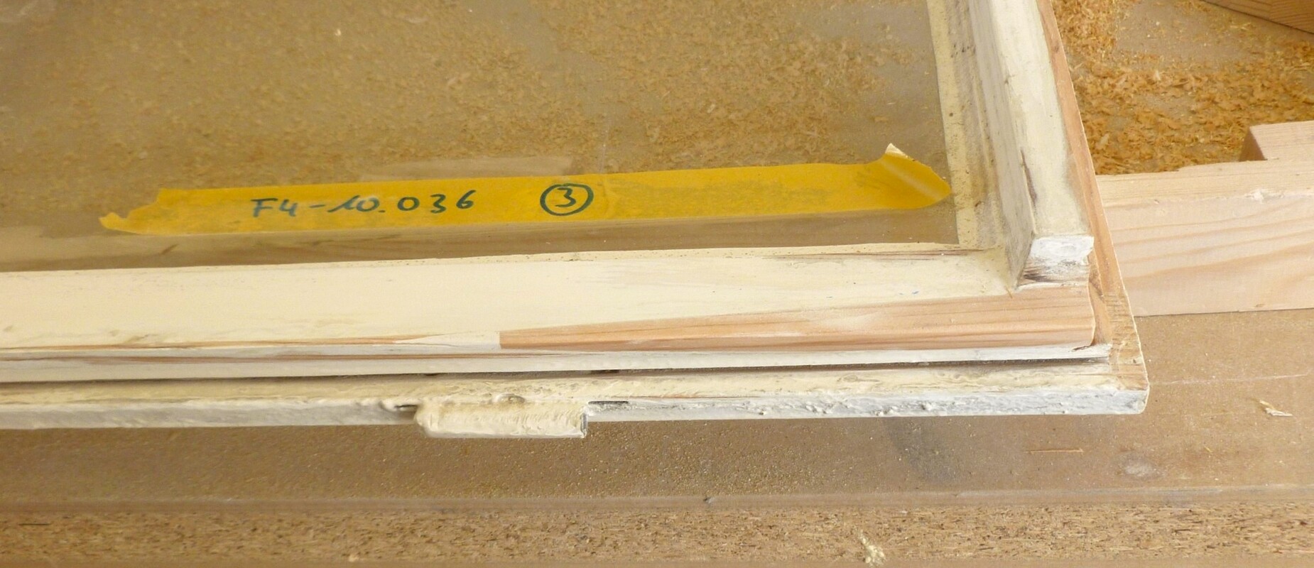 Holzfenster mit gr&uuml;nem Glas und gelbem Etikett auf einer Werkbank, umgeben von Holzsp&auml;nen.