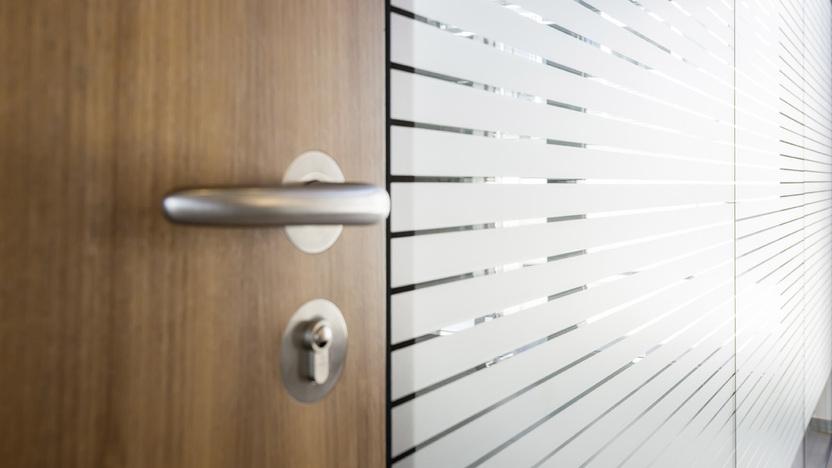 Nahaufnahme eines T&uuml;rgriffs mit Schloss an einer Holzt&uuml;r neben einer transparenter Wand mit horizontalen Streifen.