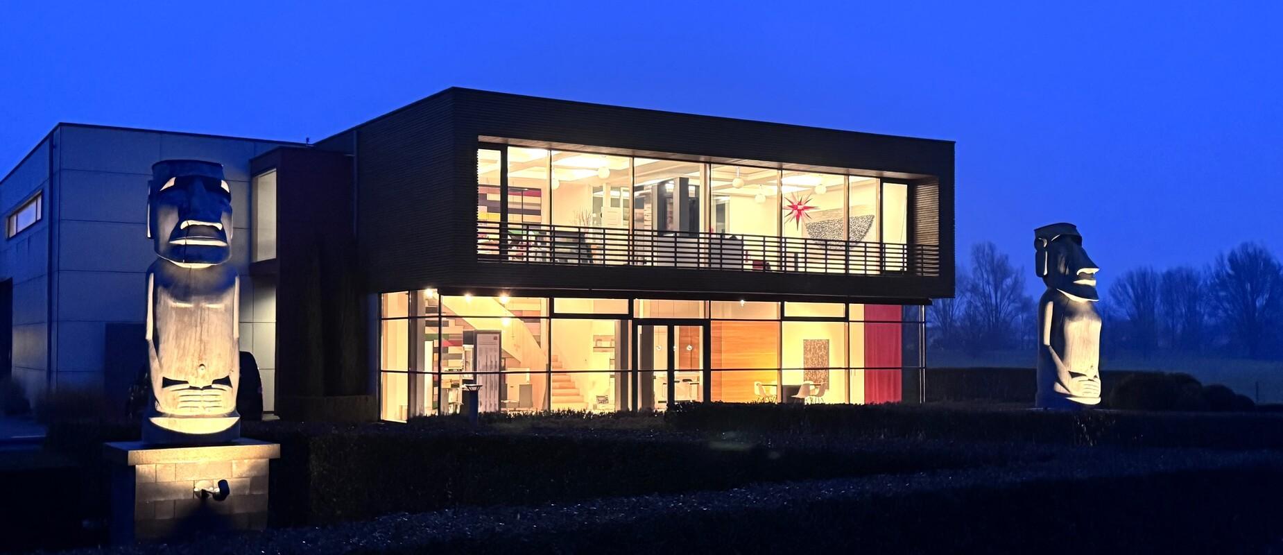 Moderne Architektur eines Geb&auml;udes bei D&auml;mmerung, flankiert von zwei Moai-Statuen in einem Garten.