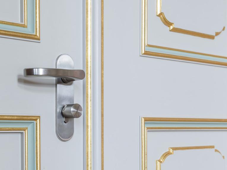 Nahaufnahme einer wei&szlig;en T&uuml;r mit goldenen Verzierungen und einer modernen T&uuml;rklinke.